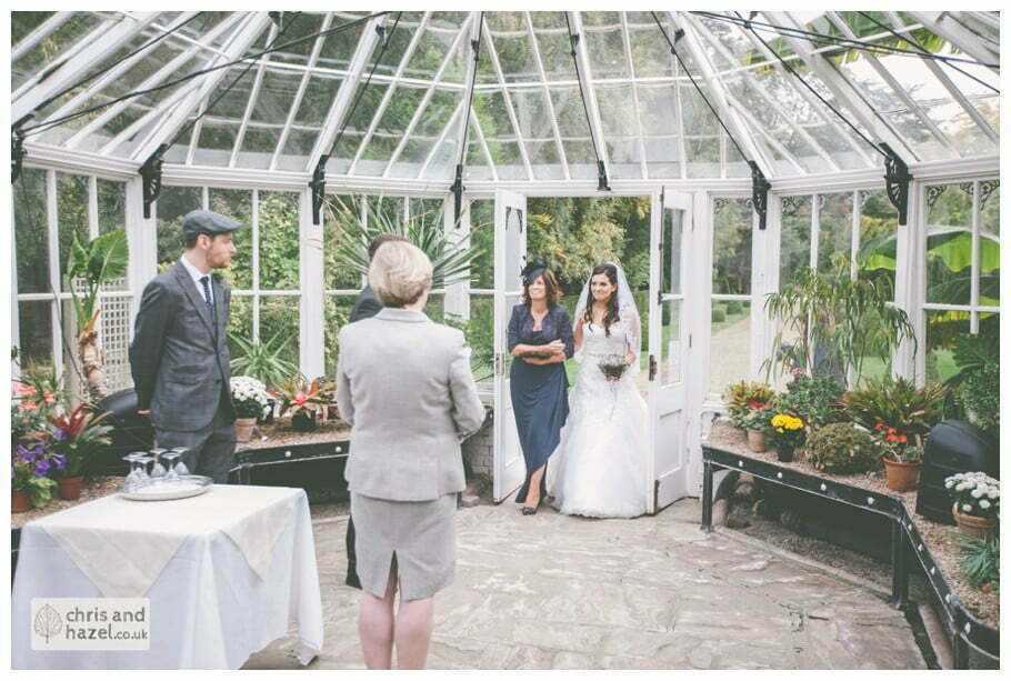 bride walking with mother of bride down aisle wedding ceremony in greenhouse conservatory documentary Hull Wedding Photographer Bishop Burton College Wedding Photography Hull by Chris and Hazel Wedding Photography Ross laurelin Matulis